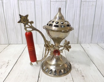 May include: A brass incense burner featuring a metallic finish, a dome-shaped lid with circular cutouts, and a decorative handle with a cylindrical red grip. The base is flared with triangular cutouts, and the structure includes leaf-shaped accents.