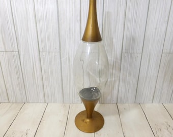 May include: A tall decorative vessel featuring a clear glass bulbous center section flanked by a metallic gold conical top and a matching circular gold base. The interior of the glass section contains a dark gray circular element at the bottom.