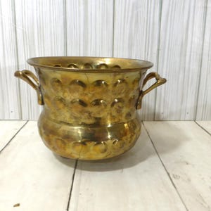 May include: A polished brass planter featuring a rounded body with a repeating hammered circular indentation pattern. The vessel includes two curved side handles and a wide, open top rim, showcasing a metallic gold finish with natural patina.