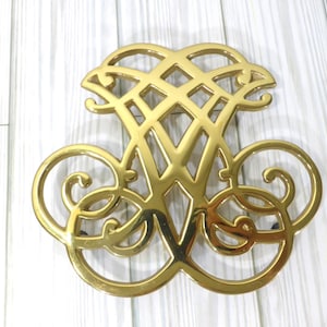 May include: A gold-colored metal trivet with an ornate design. The trivet features a stylized letter "W" in the center, surrounded by swirling patterns and a lattice-like top. The trivet is designed to protect surfaces from hot cookware.
