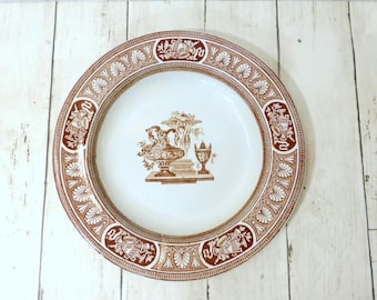 May include: A round ceramic plate featuring a white center with a brown transferware illustration of classical urns and pedestals. The wide rim is decorated with a repeating brown and white pattern of shell motifs and ornate urns.