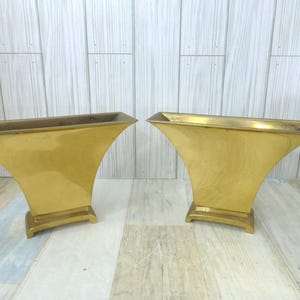 May include: Two gold-coloured metal vases with a flared, trumpet-like shape. Each vase has a rectangular top and a square base. The vases are displayed on a light-coloured wooden surface.
