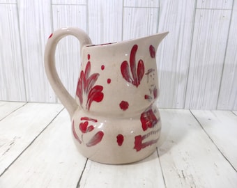 May include: A beige ceramic pitcher featuring a hand-painted red floral pattern with stylized leaves and scattered dots. The pitcher has a rounded body, a wide pouring spout, and a curved handle on the side, showcasing a glossy finish throughout.