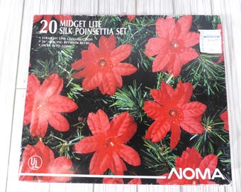 May include: A NOMA 20 Midget Lite Silk Poinsettia set in a rectangular box. The packaging features red silk poinsettia flowers with green pine needles, 20 inch spacing between bulbs, and a total length of over 14 feet. The UL logo is visible.