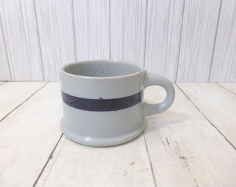 May include: A light gray ceramic mug featuring a single horizontal dark blue stripe encircling the center. The mug has a thick, sturdy handle and a slightly flared base, with a speckled texture visible throughout the light gray glazed surface.