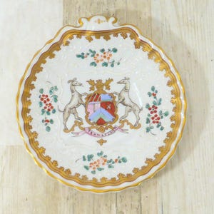 Könnte beinhalten: Ein weißer Teller mit gewelltem Rand, goldenem Rand und aufwendigem Design. Der Teller zeigt ein zentrales Wappen mit zwei Windhunden, einem Schild und floralen Akzenten in Rot, Orange und Grün. Der Teller hat einen Durchmesser von ca. 15 cm.