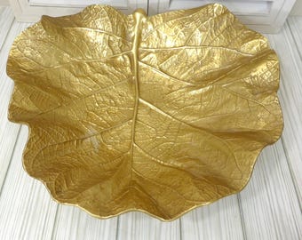 May include: A gold-colored decorative bowl shaped like a large, broad leaf. The surface features a highly detailed, embossed texture that mimics the natural veins and organic contours of a real leaf, with a metallic finish and scalloped edges.