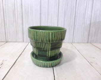 May include: A green ceramic plant pot with a matching saucer, featuring a textured exterior with alternating vertical and horizontal embossed lines. The pot has a tapered cylindrical shape and a deep, variegated forest green glaze finish.
