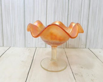 May include: A footed glass bowl featuring an iridescent orange finish and a ruffled, scalloped rim. The bowl is supported by a clear, cylindrical stem and a circular base, showcasing a reflective, metallic sheen across its curved surface.