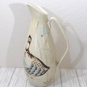 Puede incluir: Una jarra de cerámica alta de color crema con acabado moteado y asa curva. El cuerpo presenta una ilustración pintada de un pájaro con plumaje marrón, azul y negro, junto con un pequeño motivo de hoja azul cerca de la zona del pico superior.