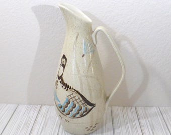 May include: A tall, cream-colored ceramic pitcher with a speckled finish and a curved handle. The body features a painted illustration of a bird with brown, blue, and black plumage, along with a small blue leaf motif near the top spout area.
