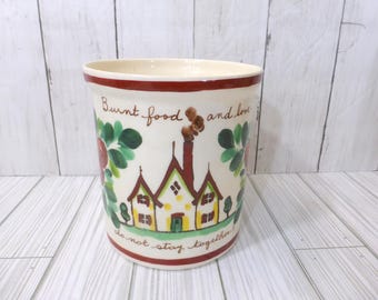 May include: A cream-colored ceramic utensil crock featuring a hand-painted illustration of a house with a smoking chimney, green foliage, and red accents. The text reads Burnt food and love do not stay together in brown script.