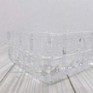 May include: A clear, square glass bowl featuring a geometric grid pattern of raised, textured squares across its exterior surface. The bowl has thick, transparent walls and a structured, faceted design that catches light, creating a repeating pattern.