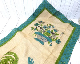 May include: A rectangular beige kitchen towel featuring a central floral arrangement in shades of blue, green, and brown. The towel is bordered by a wide teal band decorated with a small repeating star pattern and a thin green trim along the edge.