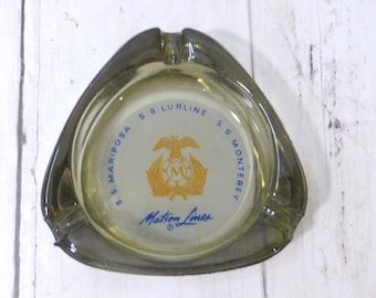 May include: A triangular olive green glass ashtray featuring a central white circular emblem with a gold eagle and the letter M. The text S.S. LURLINE, S.S. MONTEREY, S.S. MARIPOSA, and Matson Lines is printed in blue around the center graphic.