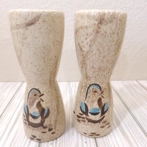 Puede incluir: Un par de hueveras de cerámica con forma de reloj de arena y acabado beige moteado. Cada pieza presenta una ilustración pintada a mano de un pequeño pájaro con detalles en azul y marrón cerca de la base, sobre un fondo claro y moteado.