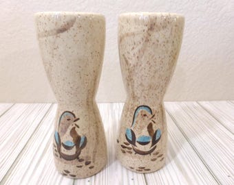 May include: A pair of tall, hourglass-shaped ceramic egg cups with a speckled beige finish. Each cup features a hand-painted illustration of a small bird with blue and brown accents near the base, set against a light, neutral speckled background.