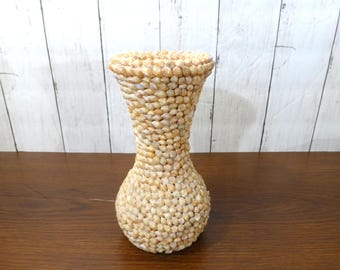 May include: A vase with an hourglass shape, entirely covered in small, light tan and cream-colored seashells. The surface features a dense, textured pattern created by the uniform arrangement of the shells, which extend from the wide base to the flared rim.