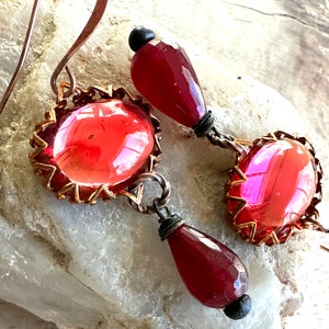 May include: A pair of dangle earrings with red glass beads and copper wire accents. The earrings feature a teardrop-shaped bead and a larger oval-shaped bead with a copper wire frame.