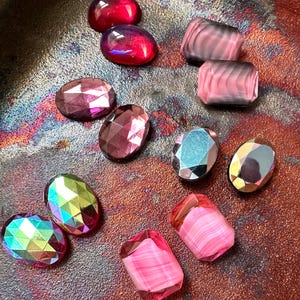 May include: A collection of faceted, colorful gemstones in various shapes. The gems include oval and rectangular cuts, with colors ranging from deep red and purple to iridescent and pink. The stones are arranged on a textured, metallic surface.