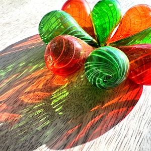 May include: A group of six glass ornaments in a teardrop shape with swirling green and orange patterns. The ornaments are arranged in a circle on a wooden surface.
