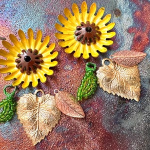 May include: A collection of vintage-style jewelry pieces, including two yellow sunflower brooches with brown centers, gold-toned leaf pendants, and green beaded grape cluster charms. The pieces are arranged on a textured, multi-colored surface.