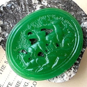May include: A vibrant green, oval-shaped carving featuring a detailed relief of deer and foliage. The jade-like piece is set against a dark, textured stone, creating a striking contrast. The carving showcases intricate details and a smooth, polished finish.