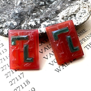 May include: Two rectangular, red-orange gemstone pendants with dark green geometric designs. The pendants have carved floral details and are displayed on a textured surface. The gemstones are likely for jewelry.