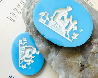 May include: Two oval-shaped bright blue cameos featuring white relief carvings of a seated figure and a smaller figure. The cameos are displayed against a textured, light-colored stone and a paper document with printed numbers and text.