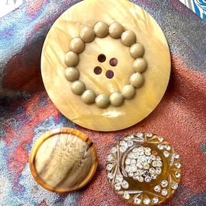 May include: A collection of three vintage buttons. The largest button is a light yellow with a ring of small, round, light brown beads around the center. The second button is a light brown with a wood grain pattern. The third button is a dark yellow with clear rhinestones.