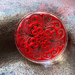 May include: A round, red decorative object with a clear, transparent border. The object features a detailed, black floral design. The design includes swirling patterns and a central flower. The object is likely made of glass or a similar material.