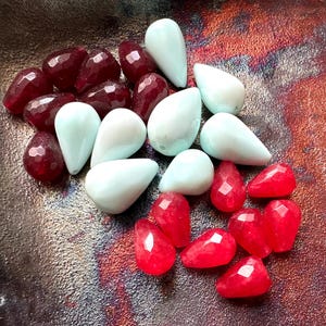 May include: A collection of gemstone beads in shades of red and light blue. The red beads have a faceted cut, while the blue beads are teardrop-shaped. The beads are arranged on a textured, multi-colored surface.