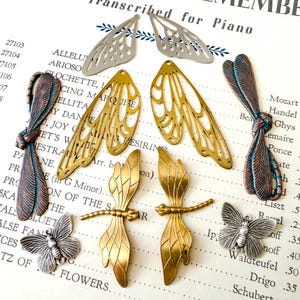 May include: A collection of metal jewelry charms including two copper-toned dragonfly bodies, two silver-toned butterfly charms, two pairs of gold-toned dragonfly wings, one pair of silver-toned dragonfly wings, and two gold-toned dragonfly pendants.