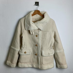 May include: Cream-coloured jacket with a faux fur collar and trim. The jacket features two front pockets, wooden buttons, and decorative buckles. The jacket is hanging on a wooden hanger against a white wall.