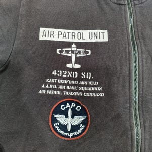Puede incluir: Primer plano de una sudadera con cremallera gris oscuro con texto y parches blancos. El texto dice "AIR PATROL UNIT", "432ND SQ." y otras frases relacionadas con el ej&eacute;rcito. Un parche circular presenta un dise&ntilde;o alado y la palabra "Encampment".