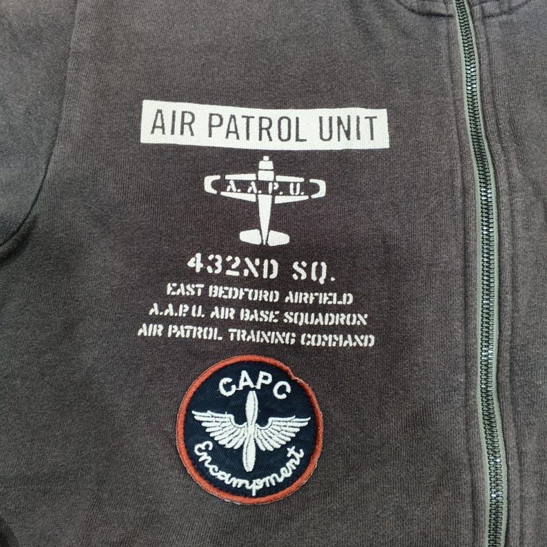 Puede incluir: Primer plano de una sudadera con cremallera gris oscuro con texto y parches blancos. El texto dice "AIR PATROL UNIT", "432ND SQ." y otras frases relacionadas con el ej&eacute;rcito. Un parche circular presenta un dise&ntilde;o alado y la palabra "Encampment".