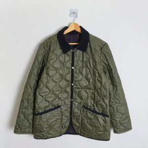 Puede incluir: Chaqueta acolchada verde oliva con cuello y ribetes de pana negra. La chaqueta tiene dos bolsillos delanteros y cierre de botones. El forro interior presenta un estampado de cuadros. La chaqueta está colgada en una percha de madera.