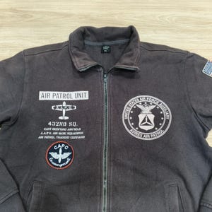 Puede incluir: Chaqueta marr&oacute;n oscuro con cremallera y varios parches y texto. La chaqueta presenta el texto "AIR PATROL UNIT" y "432ND SQ." junto con otros textos y parches. Tiene una cremallera completa y un peque&ntilde;o parche de la bandera americana en la manga derecha.