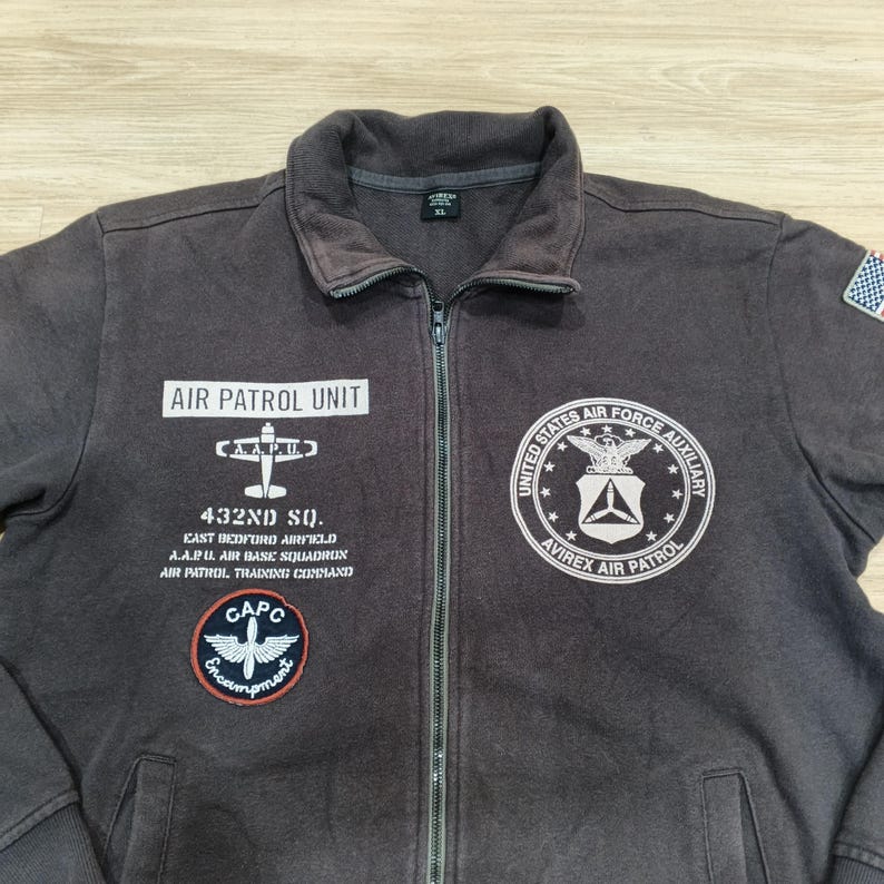Puede incluir: Chaqueta marr&oacute;n oscuro con cremallera y varios parches y texto. La chaqueta presenta el texto "AIR PATROL UNIT" y "432ND SQ." junto con otros textos y parches. Tiene una cremallera completa y un peque&ntilde;o parche de la bandera americana en la manga derecha.