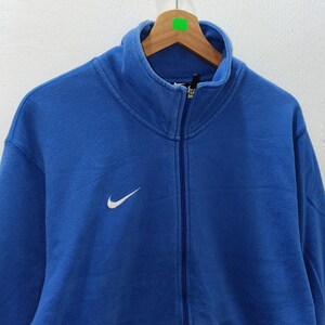 Pu&ograve; includere: Felpa blu con zip e logo Nike bianco sul petto. La felpa ha un collo alto e una cerniera a tutta lunghezza. L'indumento &egrave; appeso a una gruccia di legno.