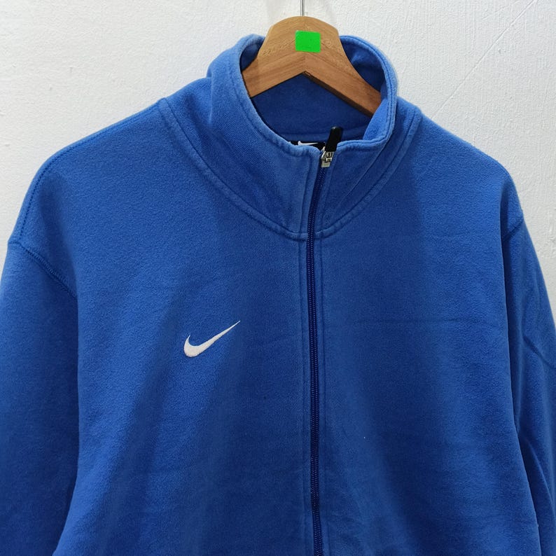 Pu&ograve; includere: Felpa blu con zip e logo Nike bianco sul petto. La felpa ha un collo alto e una cerniera a tutta lunghezza. L'indumento &egrave; appeso a una gruccia di legno.