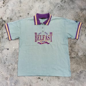Könnte beinhalten: Hellblaues Poloshirt mit lila Kragen und gelben Streifen. Der Schriftzug "DRY DOG BELFAST" ist lila. Die Ärmel haben gelbe und lila Streifen. Das Shirt hat drei Knöpfe.