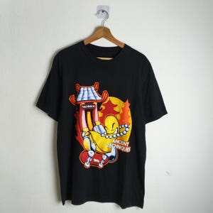 May include: Black t-shirt featuring a colorful graphic of a yellow character on a skateboard, with a pagoda and a rising sun in the background. The text "TECHDECK" is visible on the pagoda, and "ANCIENT TECHNIQUES" is written below the character.