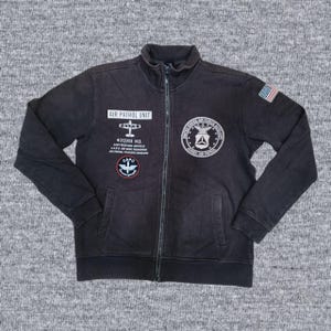 Puede incluir: Chaqueta negra con cremallera con m&uacute;ltiples parches y texto. La chaqueta presenta un parche de la bandera estadounidense y varios parches de tem&aacute;tica militar con texto como "AIR PATROL UNIT" y "UNITED STATES AIR FORCE AUTHORITY".