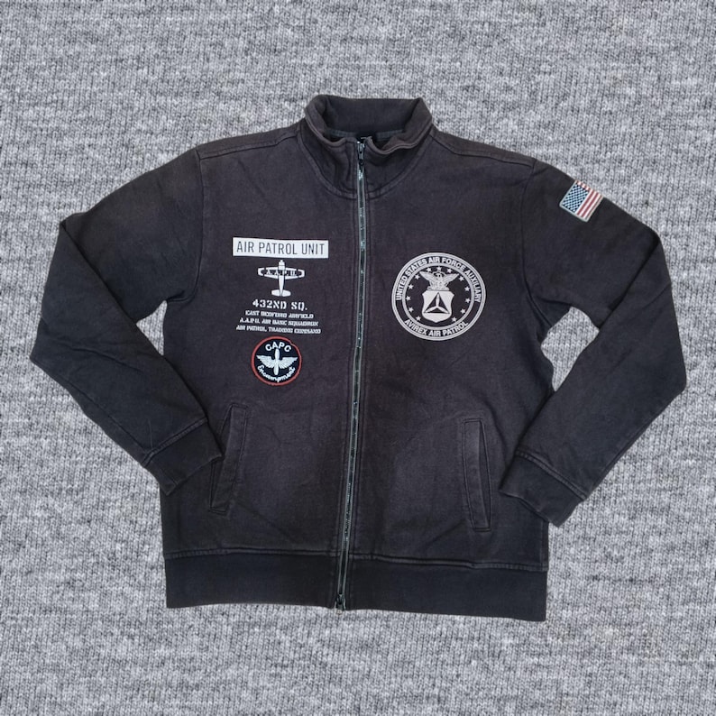 Puede incluir: Chaqueta negra con cremallera con m&uacute;ltiples parches y texto. La chaqueta presenta un parche de la bandera estadounidense y varios parches de tem&aacute;tica militar con texto como "AIR PATROL UNIT" y "UNITED STATES AIR FORCE AUTHORITY".