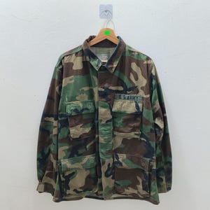 Könnte beinhalten: Tarnmusterjacke mit mehreren Taschen und dem Text "U.S. ARMY" auf der rechten Brust. Die Jacke ist eine Mischung aus grünen, braunen und schwarzen Farben. Sie hängt an einem Holzbügel.