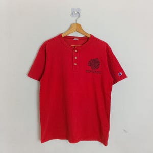 May include: Red short-sleeved shirt with a Henley neckline and three buttons. The shirt features a dark graphic of a Native American headdress and the word "NAVAJO" printed below it. A small Champion logo is on the left sleeve.