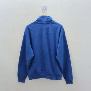 Pu&ograve; includere: Una felpa blu reale con collo alto e maniche lunghe. Il capo &egrave; appeso a una gruccia di legno contro un muro bianco. La vista posteriore della felpa mostra il suo colore pieno e il design semplice.