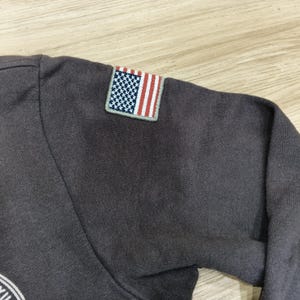 Puede incluir: Una camiseta de manga larga gris oscuro con un peque&ntilde;o parche de la bandera estadounidense en la manga superior izquierda. El parche de la bandera presenta un campo azul con estrellas blancas y rayas rojas y blancas. La camiseta es de material suave.