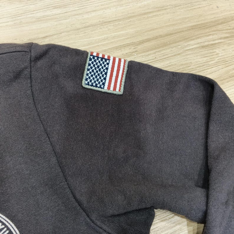 Puede incluir: Una camiseta de manga larga gris oscuro con un peque&ntilde;o parche de la bandera estadounidense en la manga superior izquierda. El parche de la bandera presenta un campo azul con estrellas blancas y rayas rojas y blancas. La camiseta es de material suave.